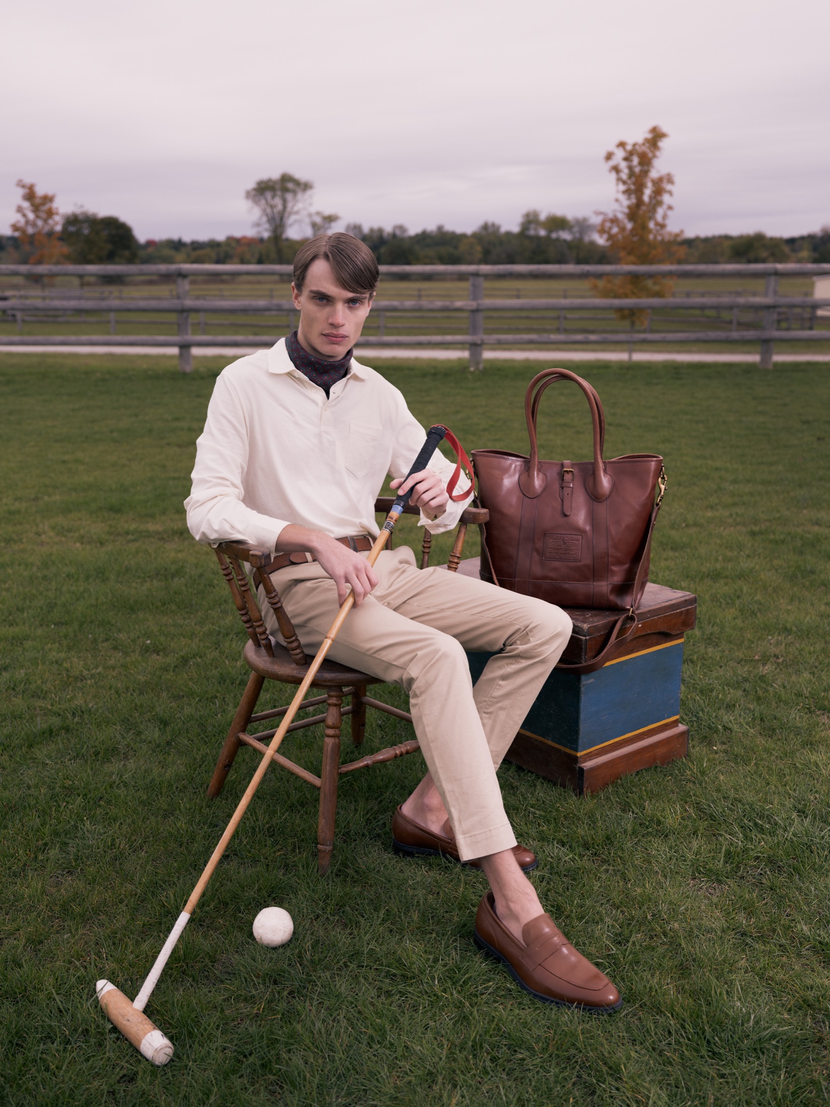 Outdoor fashion portrait of a seated model holding a mallet beside a leather bag.